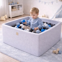 Dziecko bawiące się w jasnoszarym, kwadratowym suchym basenie z piłeczkami w kolorze niebieskim, czarnym i białym