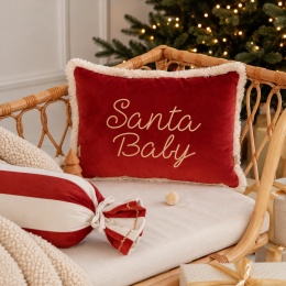 Czerwona welwetowa poduszka świąteczna Santa Baby ze złotym napisem i kremowym ozdobnym obszyciem, na rattanowej sofie przy choince.